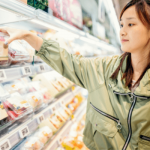 A young woman grocery shopping in the prepared foods section, reading nutrition facts and wearing a warm coat.