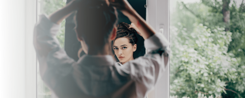 a brunette young woman looks pensive as she looks at her reflection in the mirror.