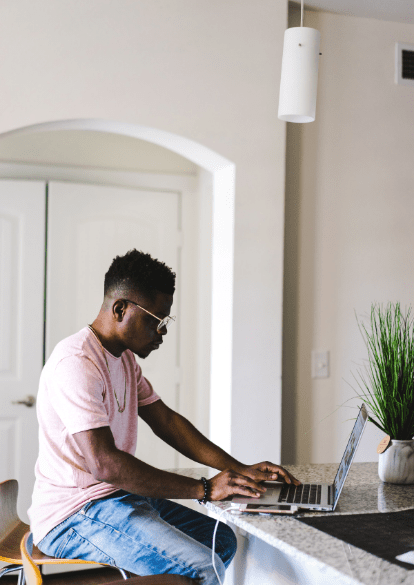 a young man wearing a pink shirt sits at his kitchen counter, participating in virtual day treatment